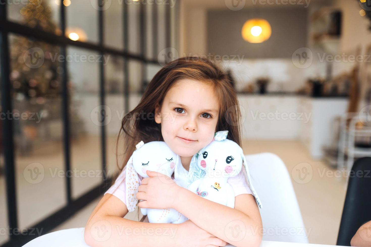 Little smiling girl hugging favorite toys while sitting at table photo