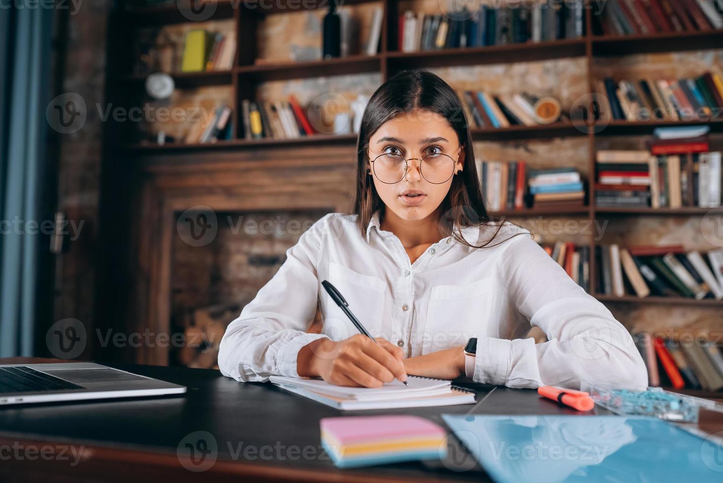 una joven con anteojos escribe en un cuaderno mientras se sienta en la mesa foto