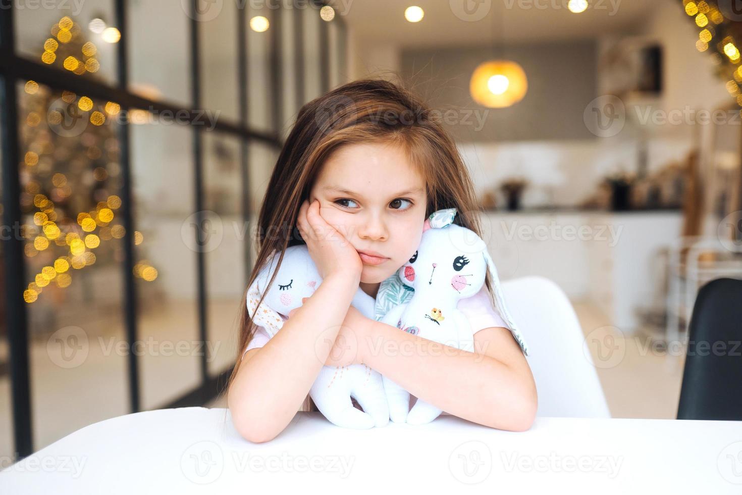 Little sad girl hugging favorite toy while sitting at table photo