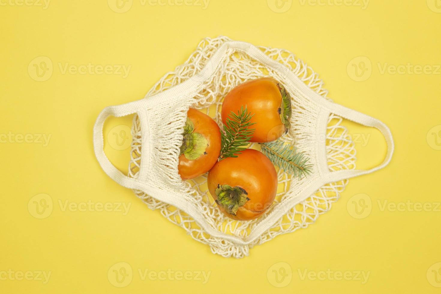 Persimmon fruits lie in the white mesh bag on a yellow background. Healthy food concept photo