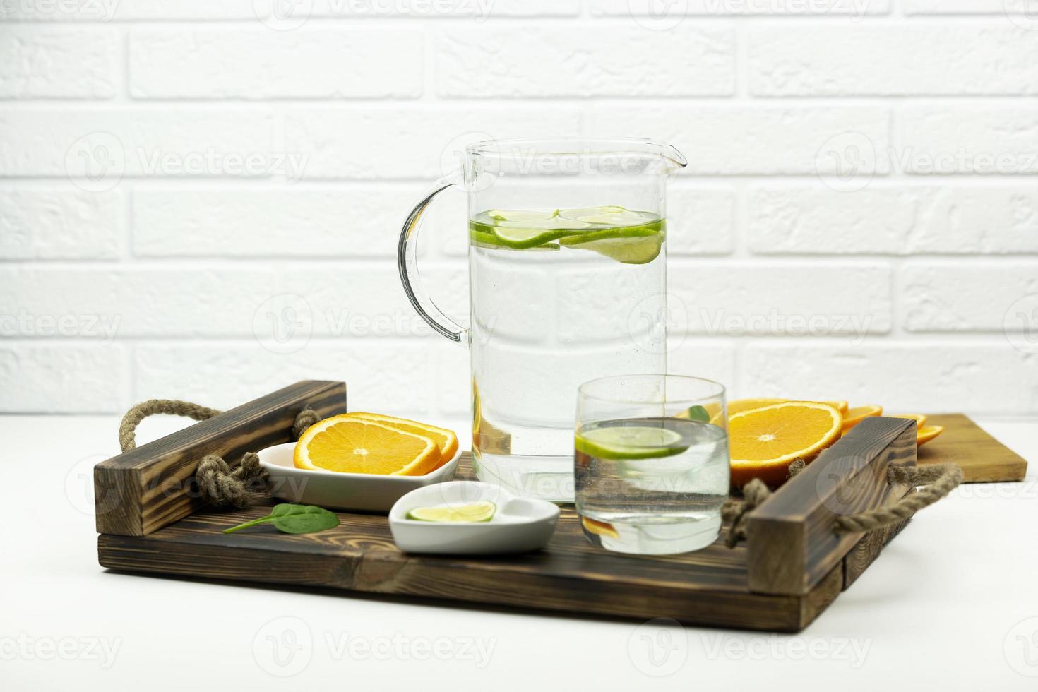 A home made lemonade made of lime stands in a glass and jug on a wooden tray with oranges around photo