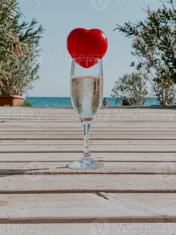 un corazón rojo se encuentra en la copa de champán con el mar y las palmeras al fondo foto