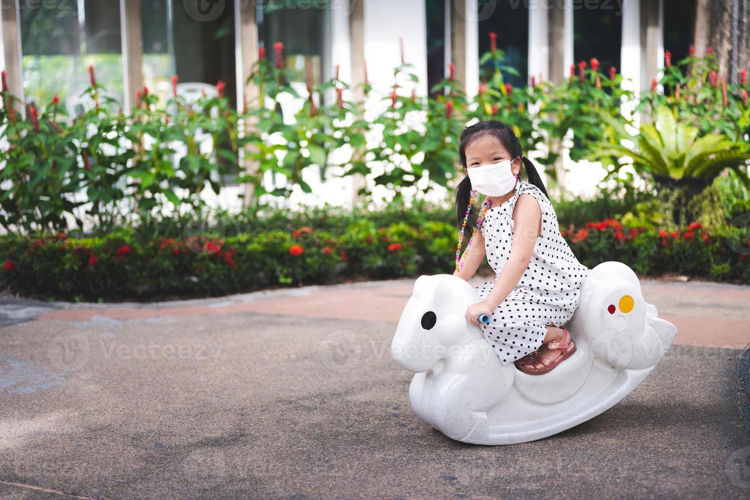 Portrait image child 5 years old. Kid wearing medical face mask to prevent toxic dust pm2.5. Little girl is having fun playing on white plastic rocking horse. Happy children smile sweetly. Empty space photo