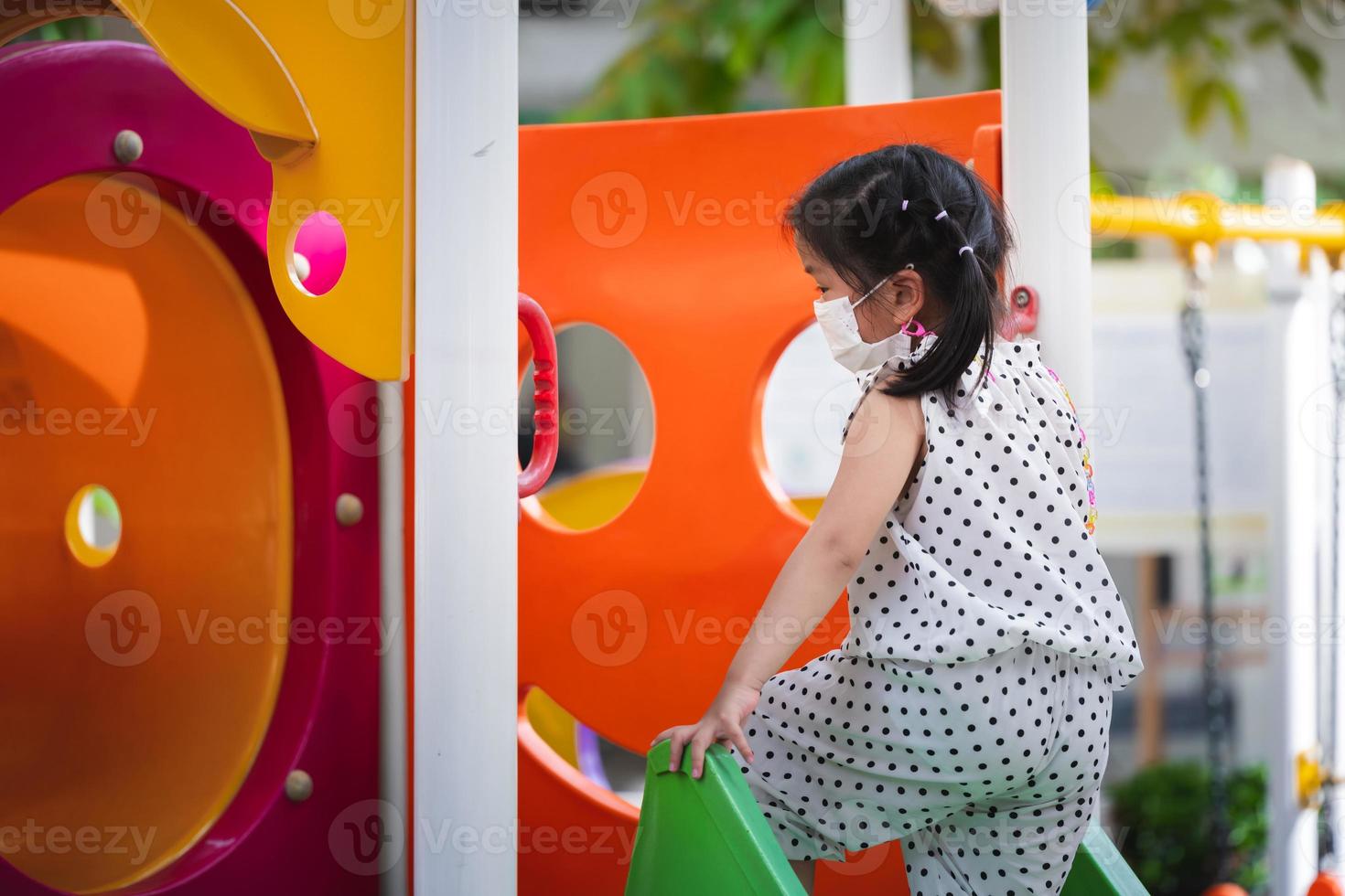 Portrait image child 5-6 years old. Cute girl wears white medical face mask while playing on slider at outdoor playground, to prevent PM2.5 dust and virus. Summer or spring time. Side view. Copy space photo