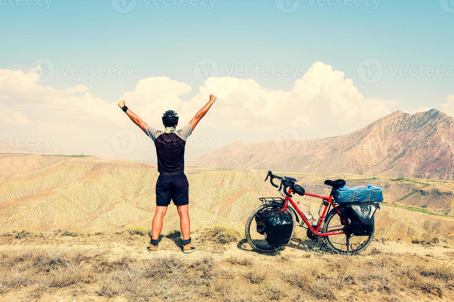 Cinematic inspirational excited fit caucasian male cyclist stand on top viewpoint by red touring bicycle in deserted mountains background with hands up confident carefree lifestyle photo