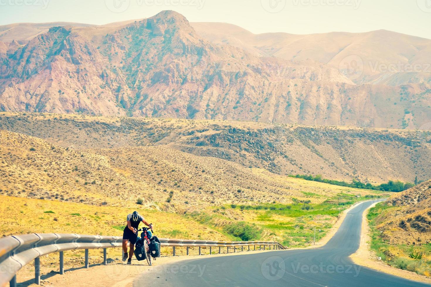 Caucasian male cyclist push his heavy touring bicycle uphill in extreme heat outdoors in mountains. Bicycle touring adventure lifestyle concept.Hard cycle uphill and Determination photo