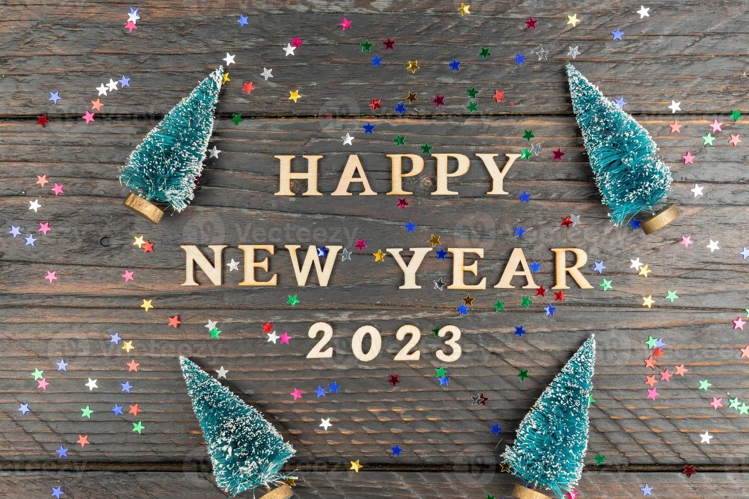 Happy New year 2023 celebration. Wooden text and numbers 2023 on wooden background with scattered multicolored confetti and fur tree miniature toys. photo