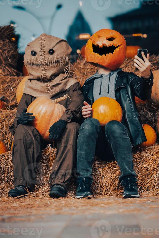 A guy with a  pumpkin head sits next to a scarecrow photo