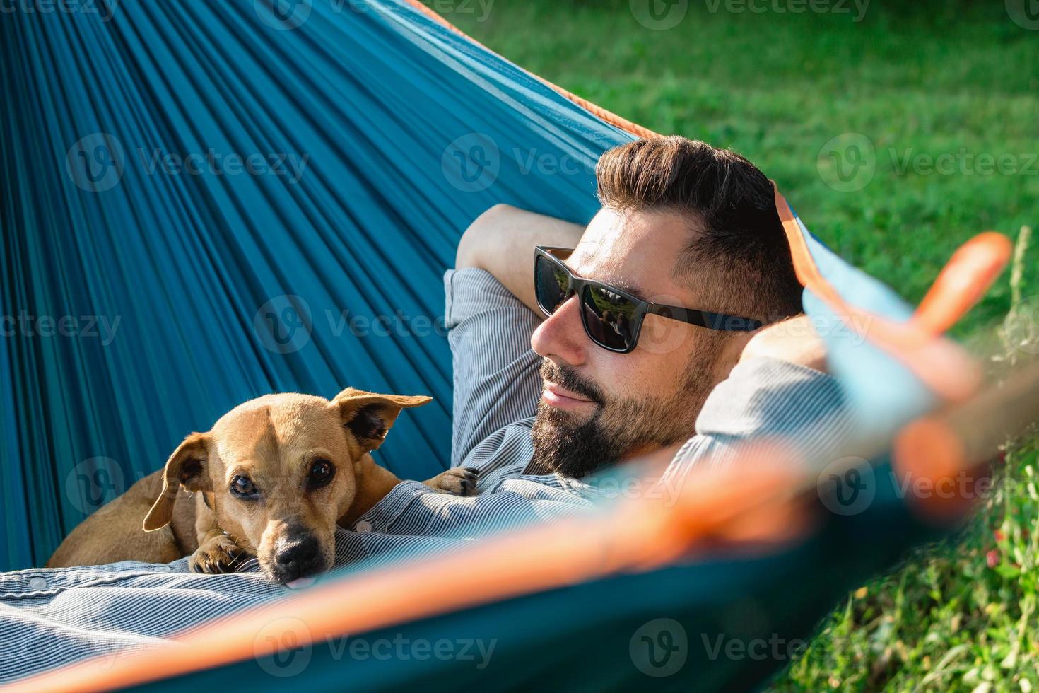 Smiling attractive European man in sunglasses is resting in hammock with his cute little dog. photo