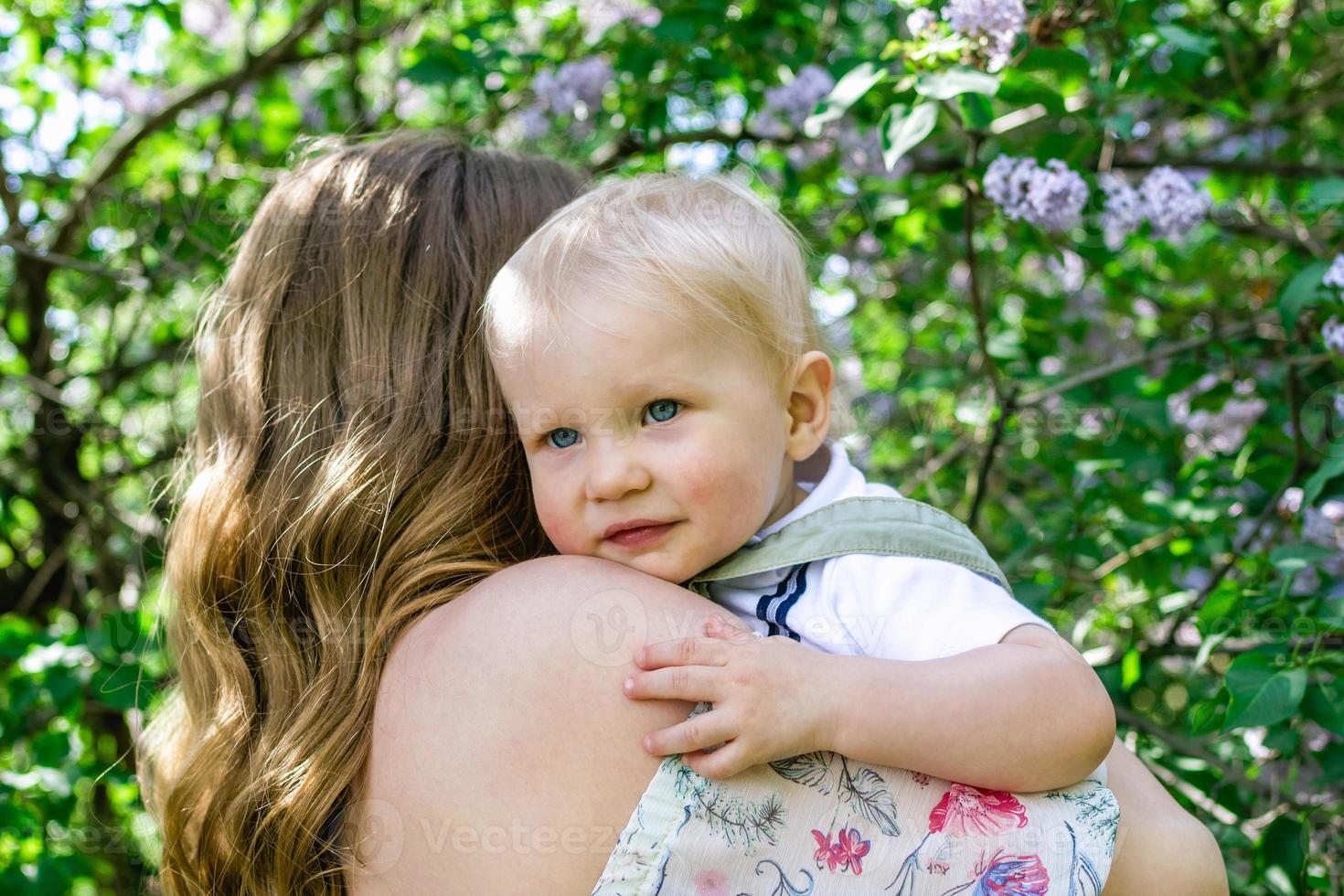 Portrait of mother with little son in her hands in park with lilac blossom tree, back view. Happy mother and child. photo