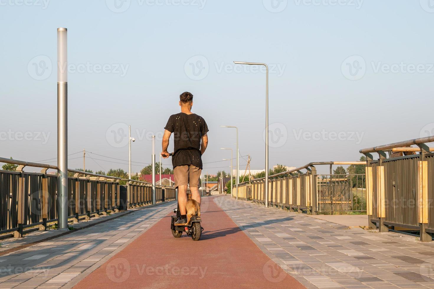 European man is riding on electric scooter with his small dog. Walking with dog on empty street of city at sunny day. photo