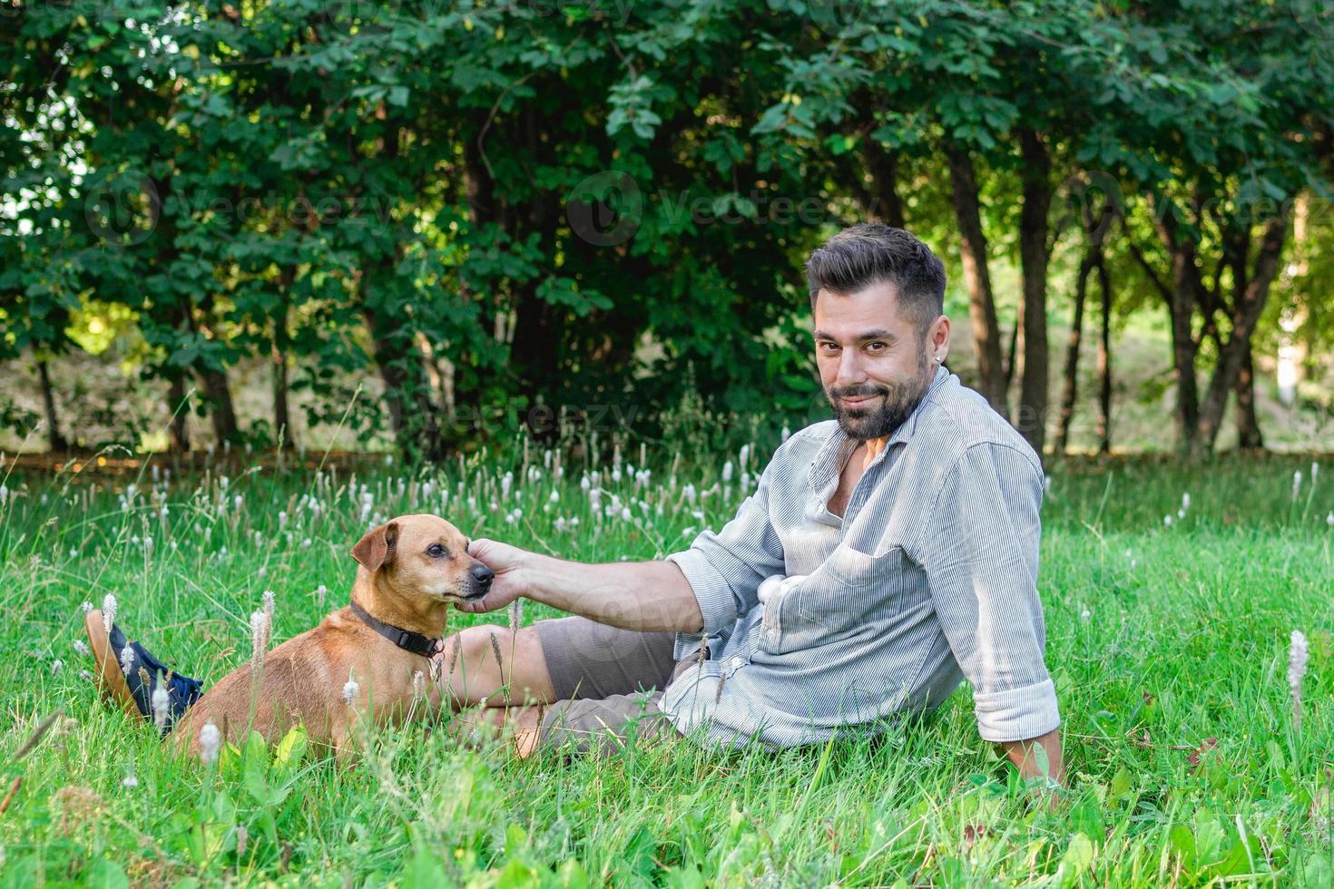 apuesto hombre sonriente sentado en la hierba con su perro en el parque. concepto de relación humana y mascota. foto