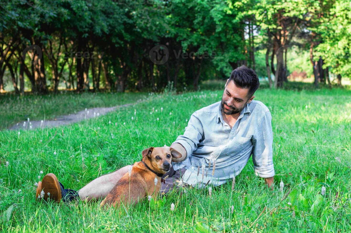 Handsome smiling man sitting on grass with his dog in park. Concept of human and pet relationship. photo