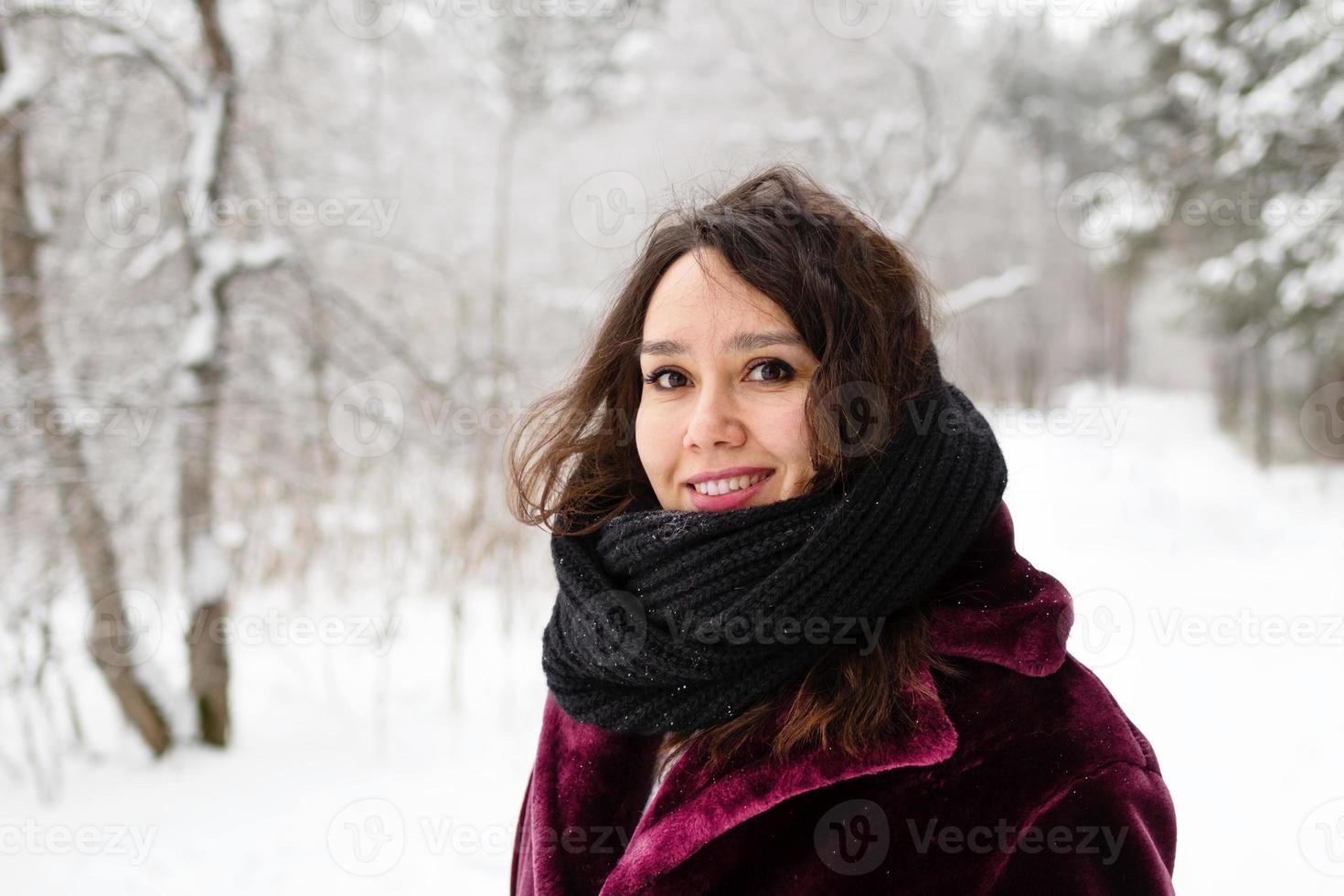Beautiful smiling woman with long brown hair in a coat from faux fur and big black scarf on a background of winter forest. photo