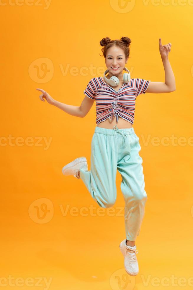 Full length photo of joyful lady jumping high listening modern youngster playlist music through cool earflaps isolated on yellow background