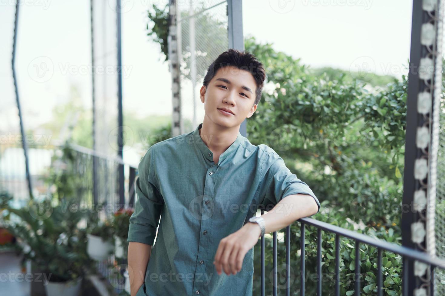 A portrait of a handsome young adult male that is leaning on a railing, with the sun shining and trees around. photo