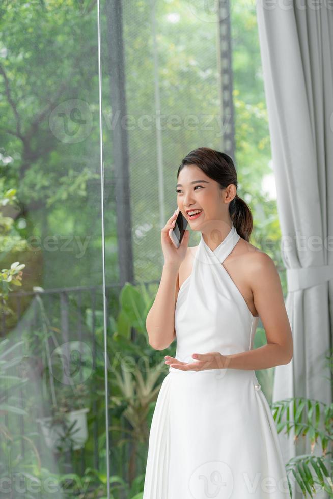 Beautiful adult woman at home living room standing by bright window talking on cellphone. photo