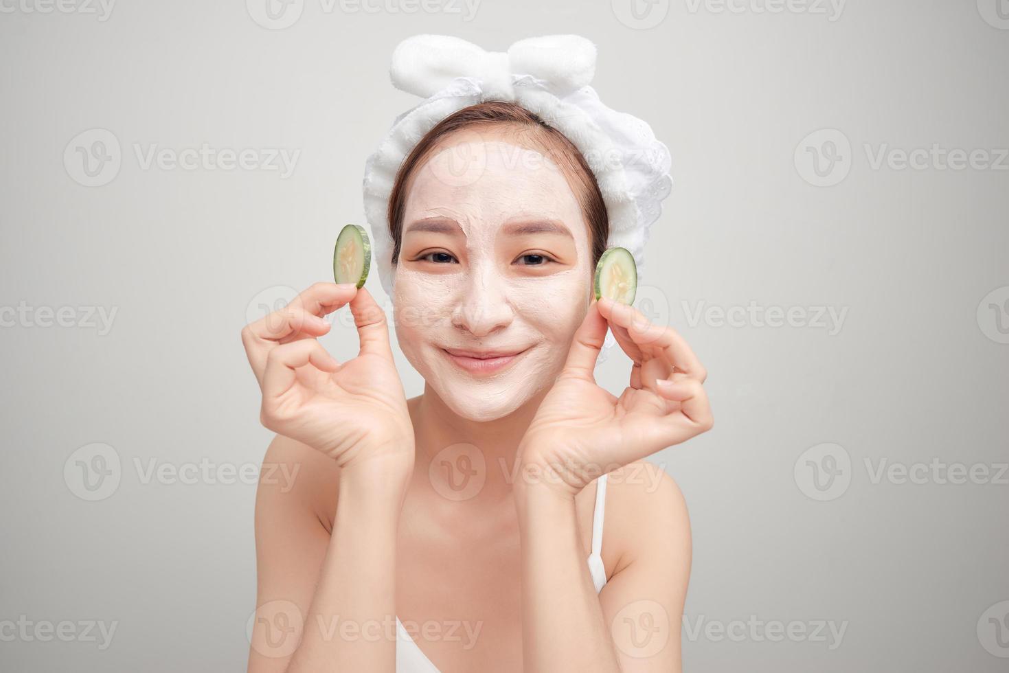 Beautiful young woman with facial mask on her face holding slices of fresh cucumber. photo