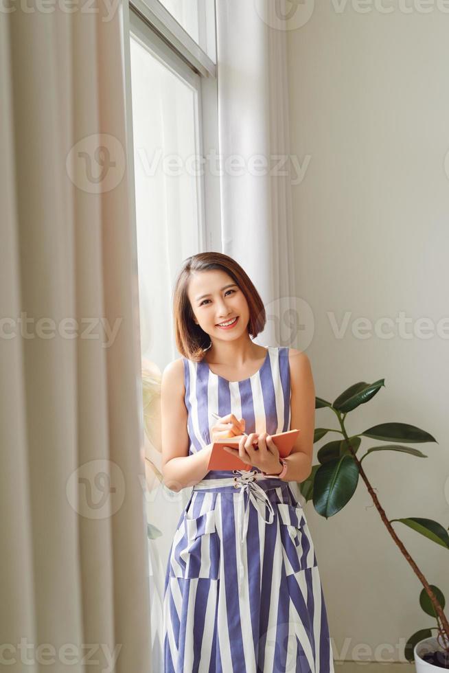 Young female standing at window in modern workspace taking notes in diary looking away with gentle smile photo