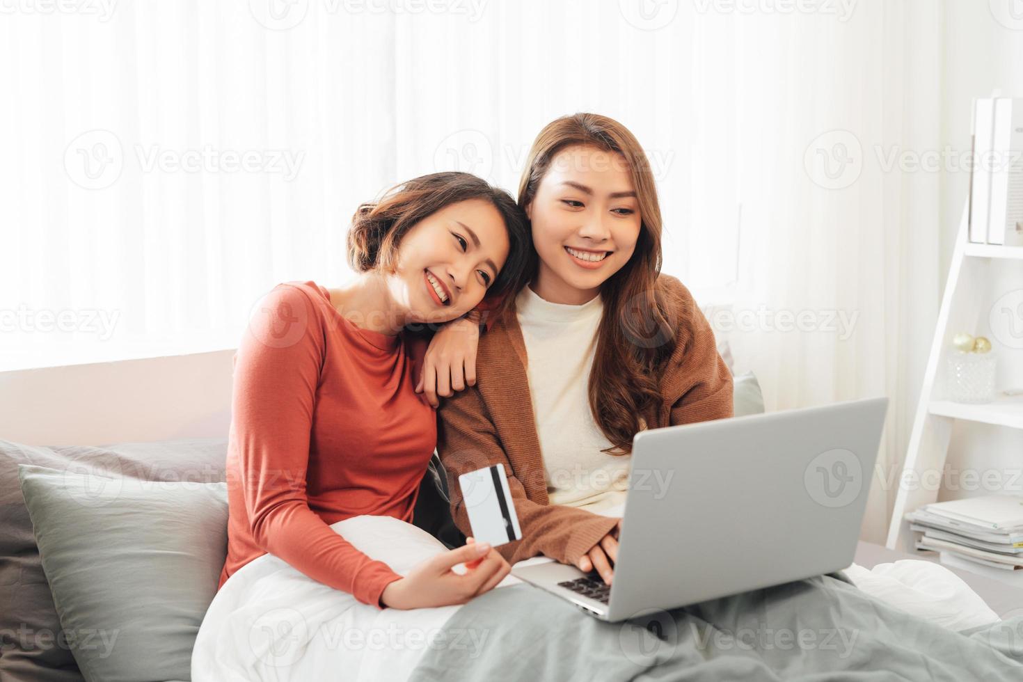 Two beautiful women lying in bed, holding a credit card and a laptop in it photo