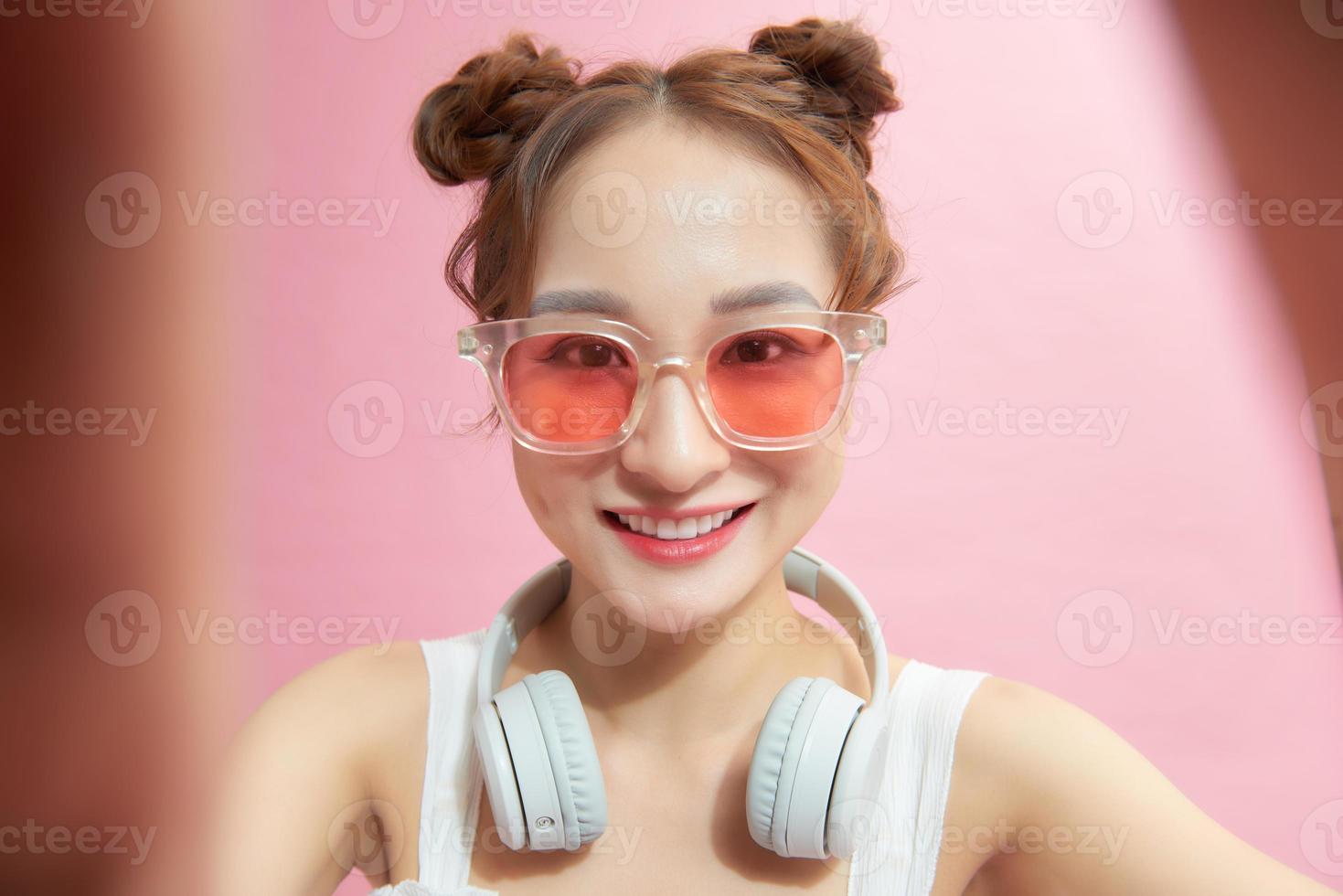 Image of pleased asian woman laughing and showing peace sign while taking selfie photo on cellphone isolated over pink background