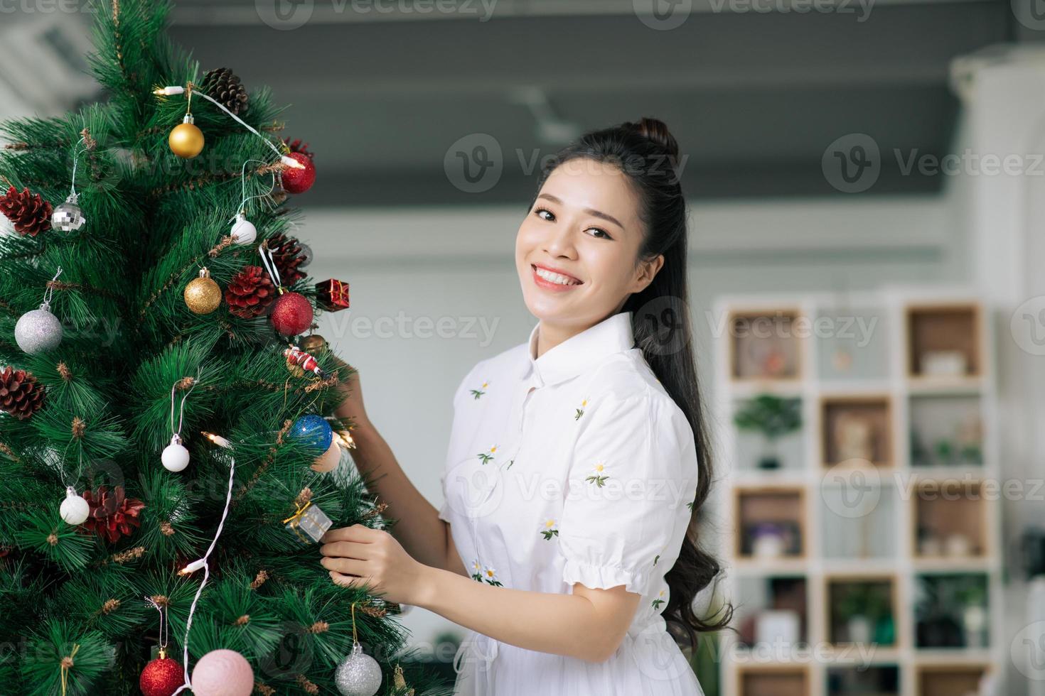 Happy beautiful young woman decorating christmas tree with red ornament at home. photo