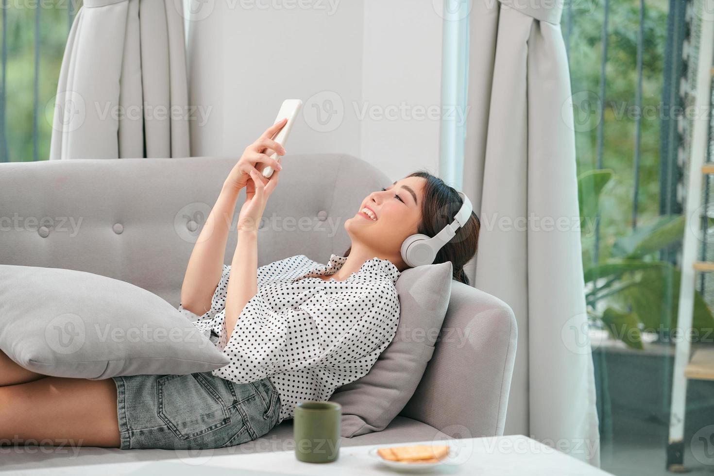 Photo of modern asian woman wearing headphones holding cell phone while lying on sofa in bright apartment