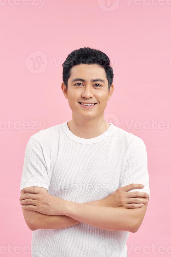 Picture of young happy asian man standing with arms crossed over pink wall. Looking at camera. photo