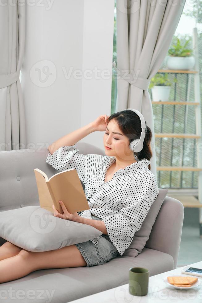 una joven asiática atractiva se relaja descansando usando un teléfono inteligente para reproducir la aplicación de transmisión de música durante la lectura de un libro sentado en un sofá o un sofá en el jardín interior de su casa foto