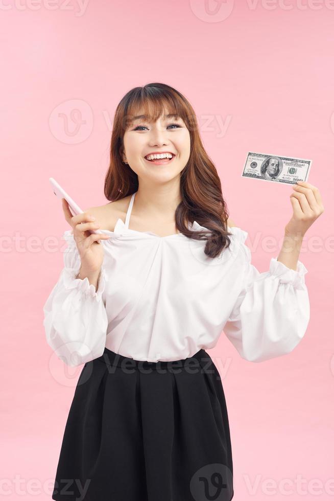 Portrait of an excited happy girl showing bunch of money banknotes while holding mobile phone and looking at camera isolated over pink background photo