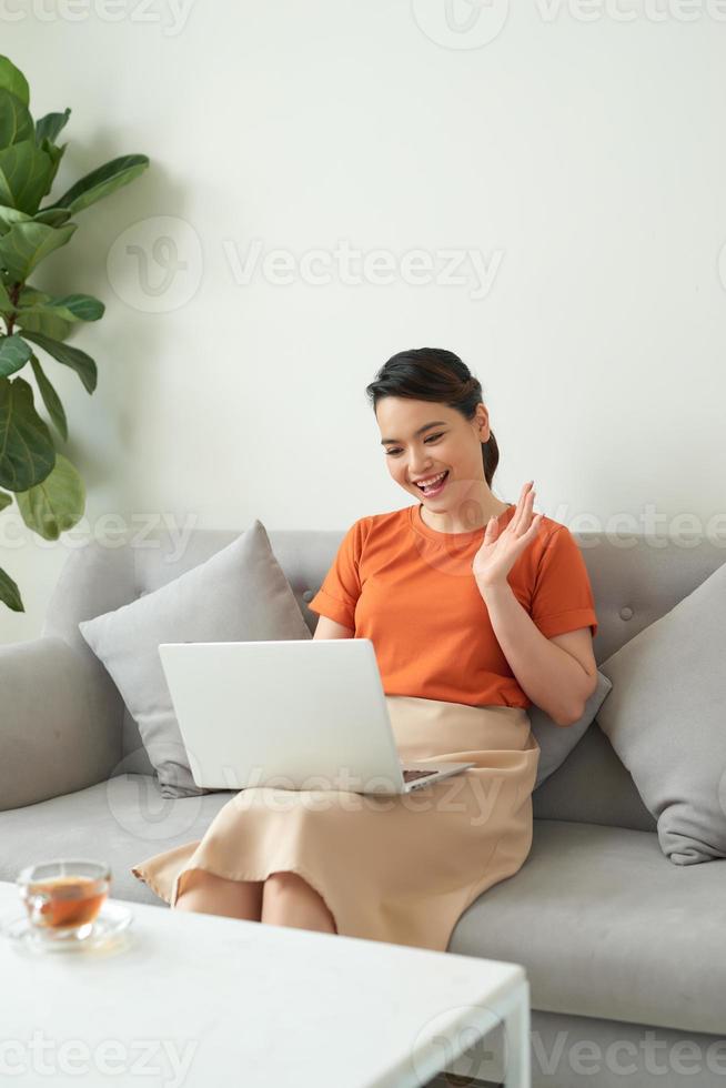 mujer asiática sonriente saludando con la mano, usando una laptop, mirando la pantalla. chatear por video en línea o estudiar en línea foto