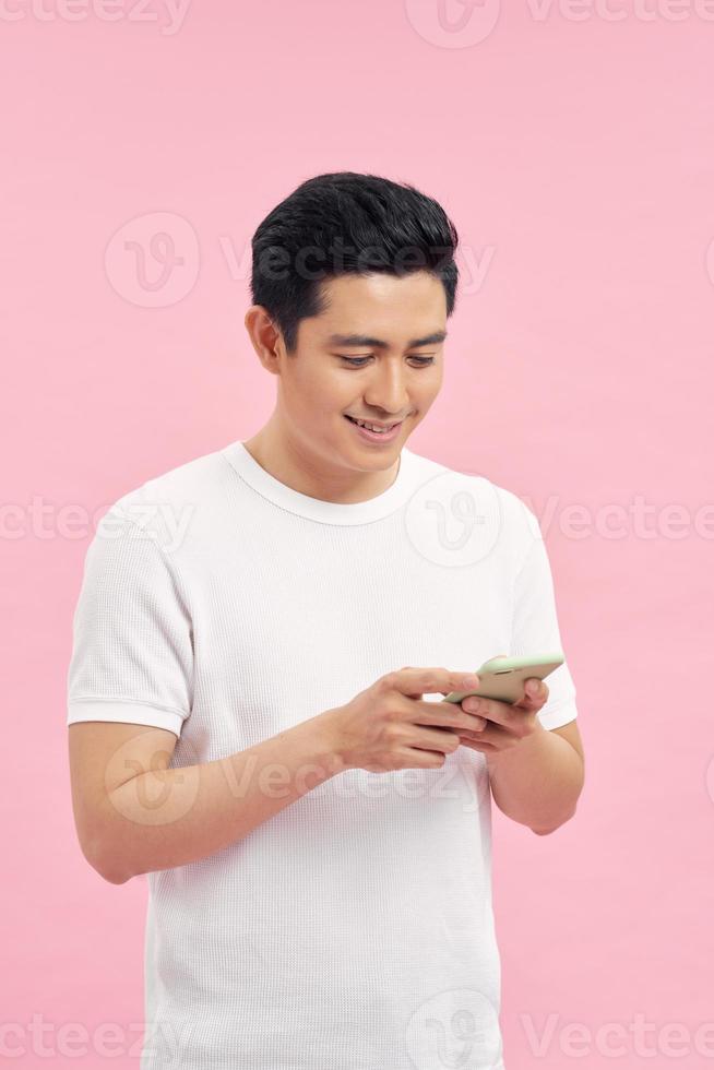 Waist-up portrait of joyful Vietnamese man leaning on gray wall while browsing Internet on smartphone, studio shot photo