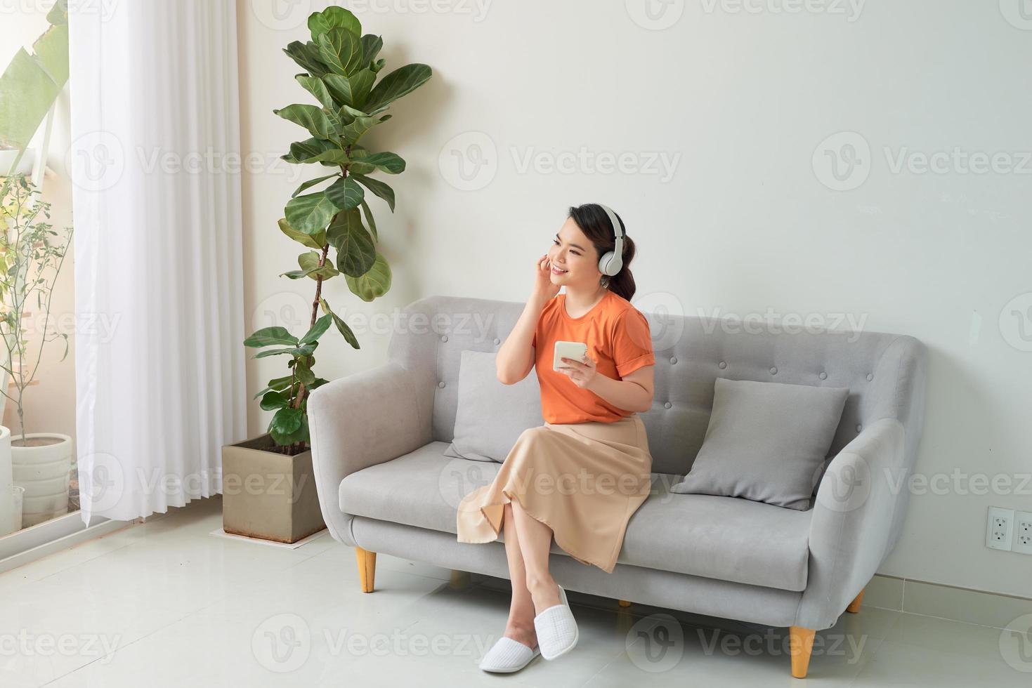 mujer joven encantada escuchando música con auriculares y usando el teléfono móvil mientras se apoya en un sofá en casa foto