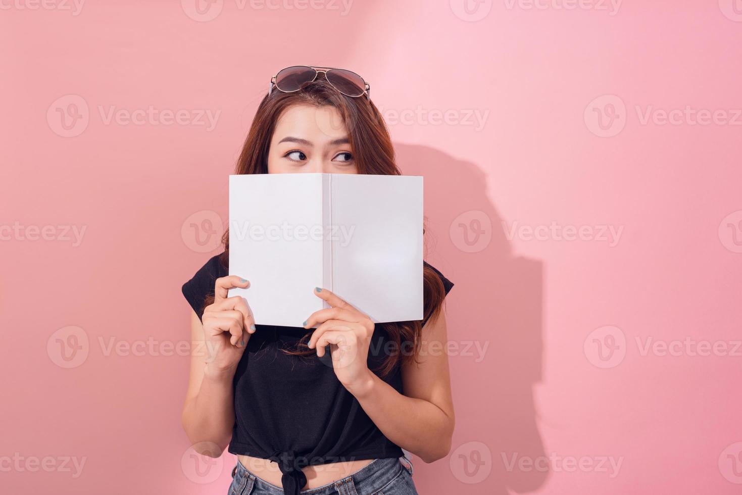 retrato de una linda joven escondida detrás de un libro abierto y mirando hacia otro lado aislada sobre el fondo de la pared rosa foto