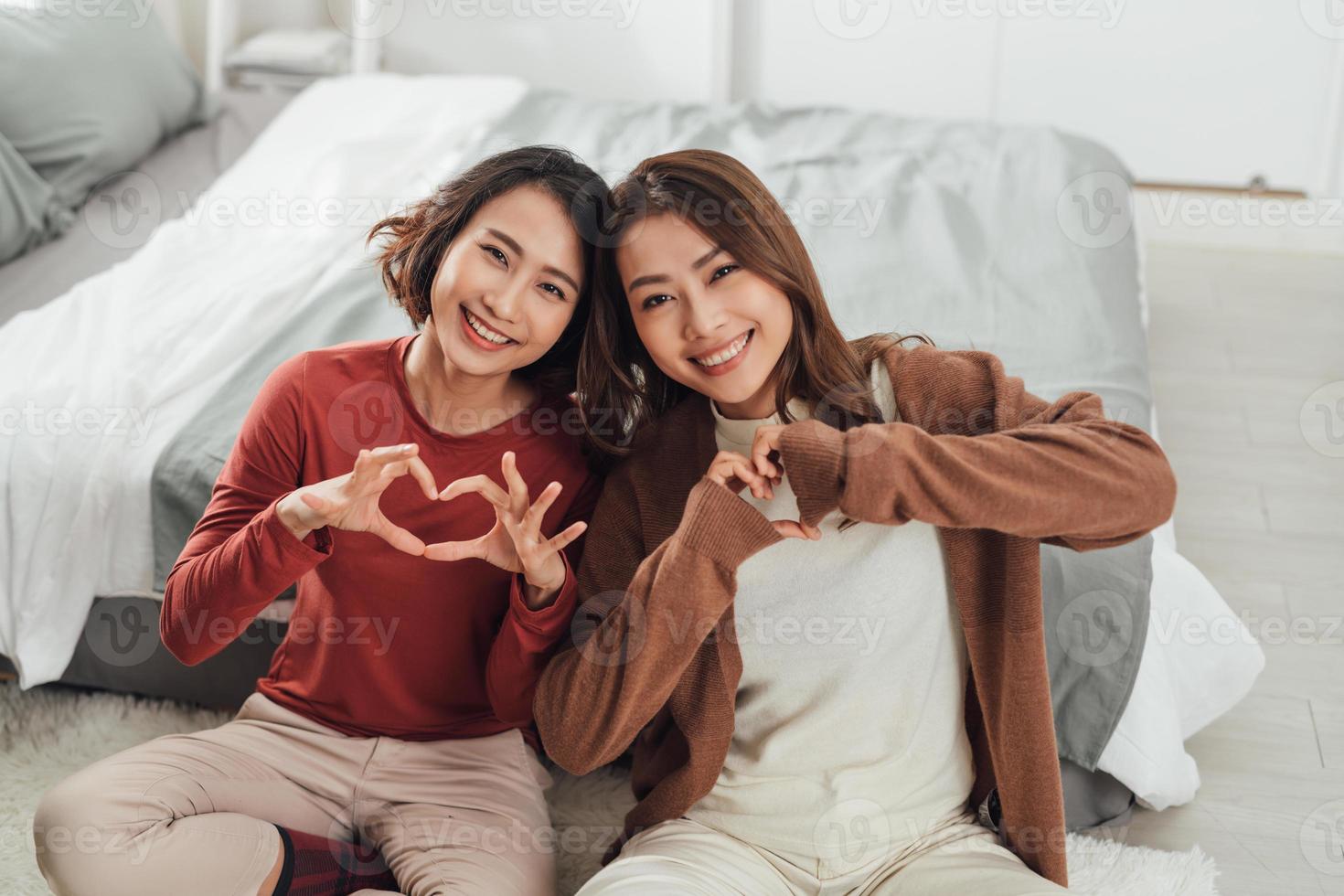 Two young women in bedroom at home lying on bed making heart shape with hands photo