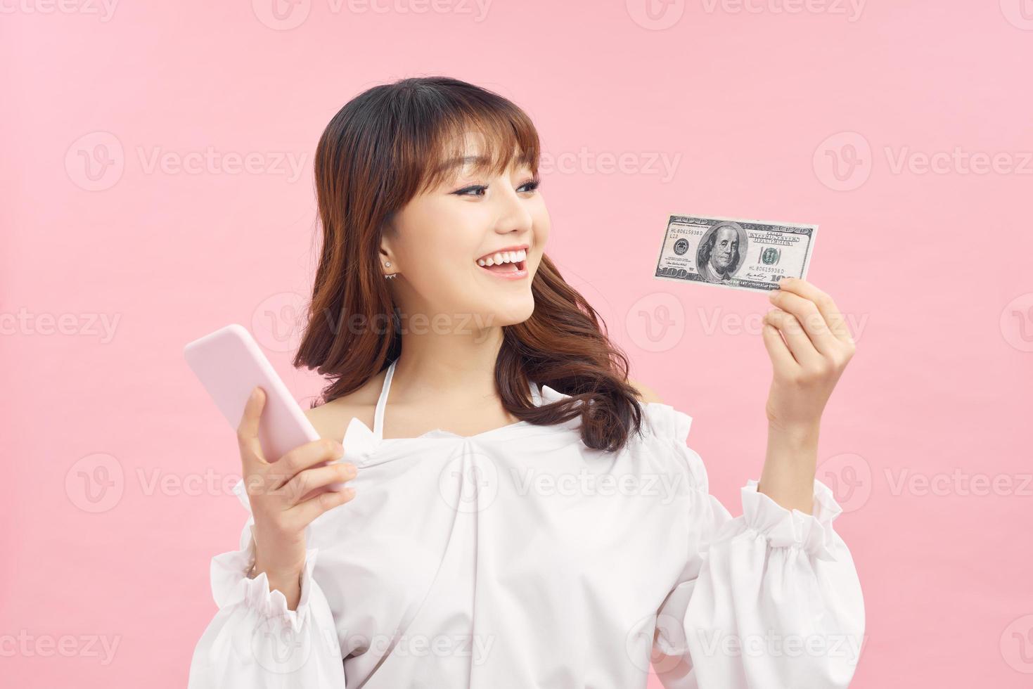 Portrait of a smiling young girl in dress standing over pink background, holding mobile phone, showing money banknotes photo