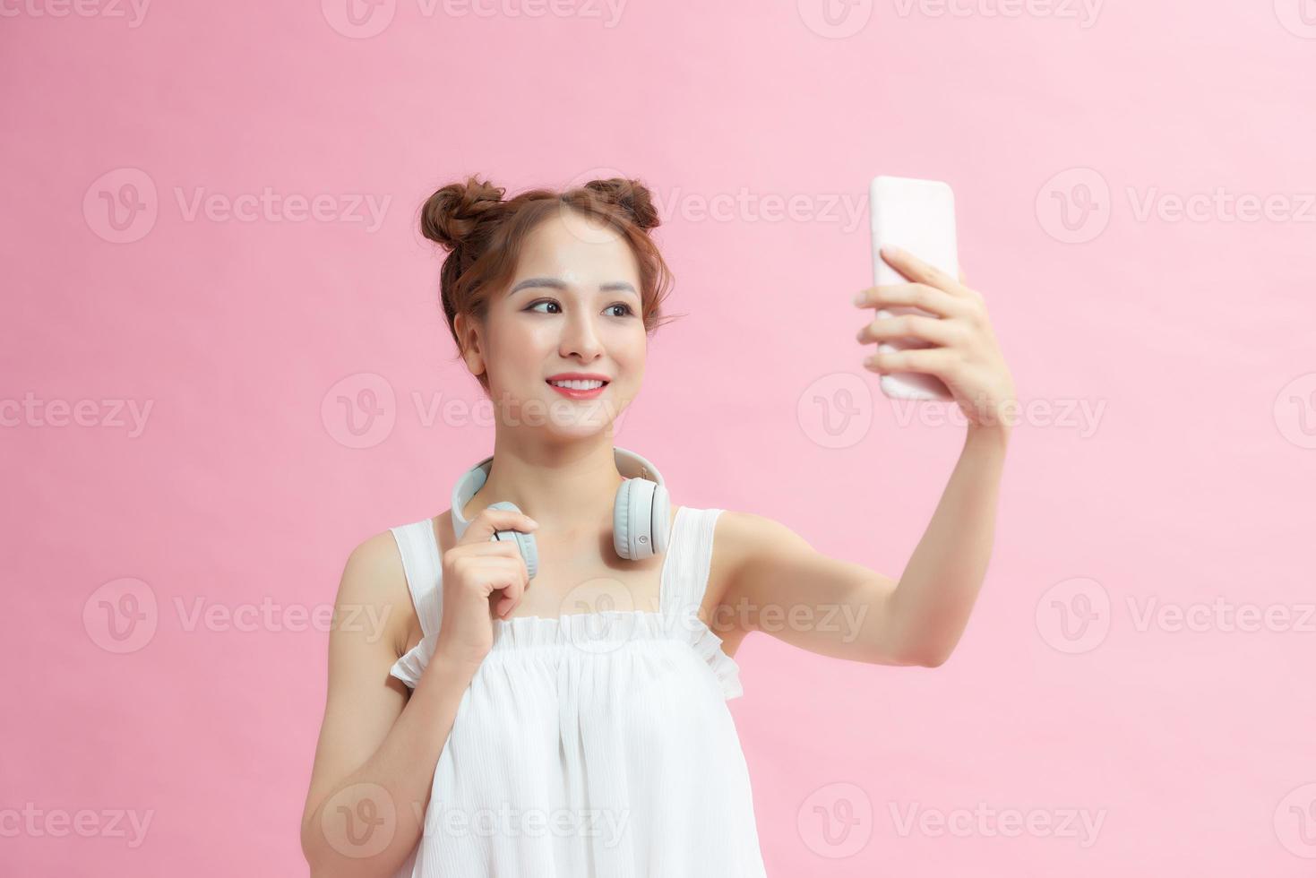 Lovely playful young woman taking selfie with mobile phone over pink background photo