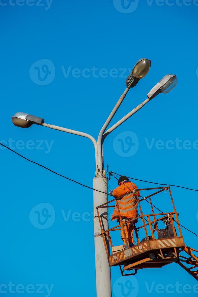A municipal worker in protective equipment performs hazardous work to eliminate an interruption in the power grid. A worker repairs a street lamp from an aerial platform basket. photo