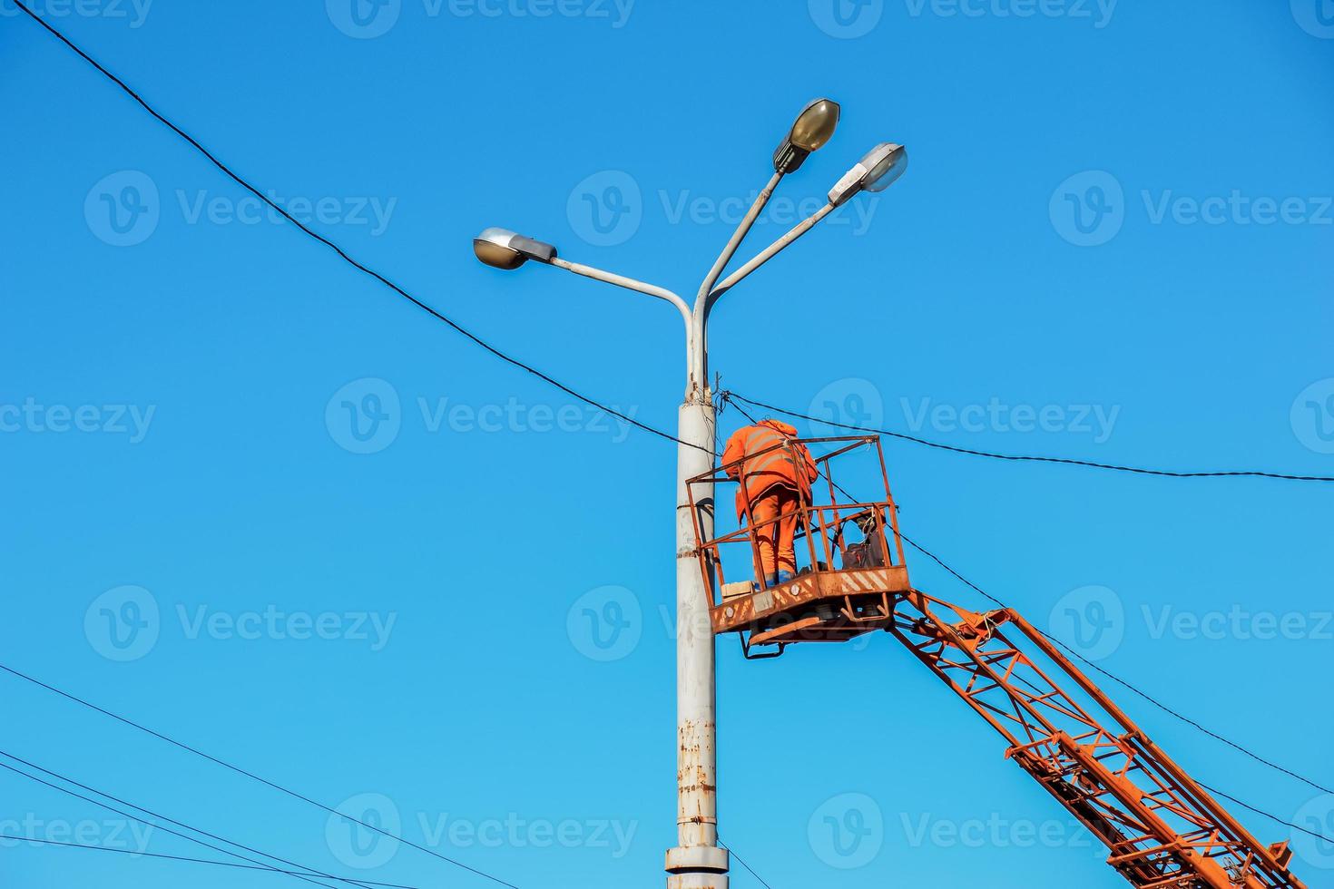 A municipal worker in protective equipment performs hazardous work to eliminate an interruption in the power grid. A worker repairs a street lamp from an aerial platform basket. photo