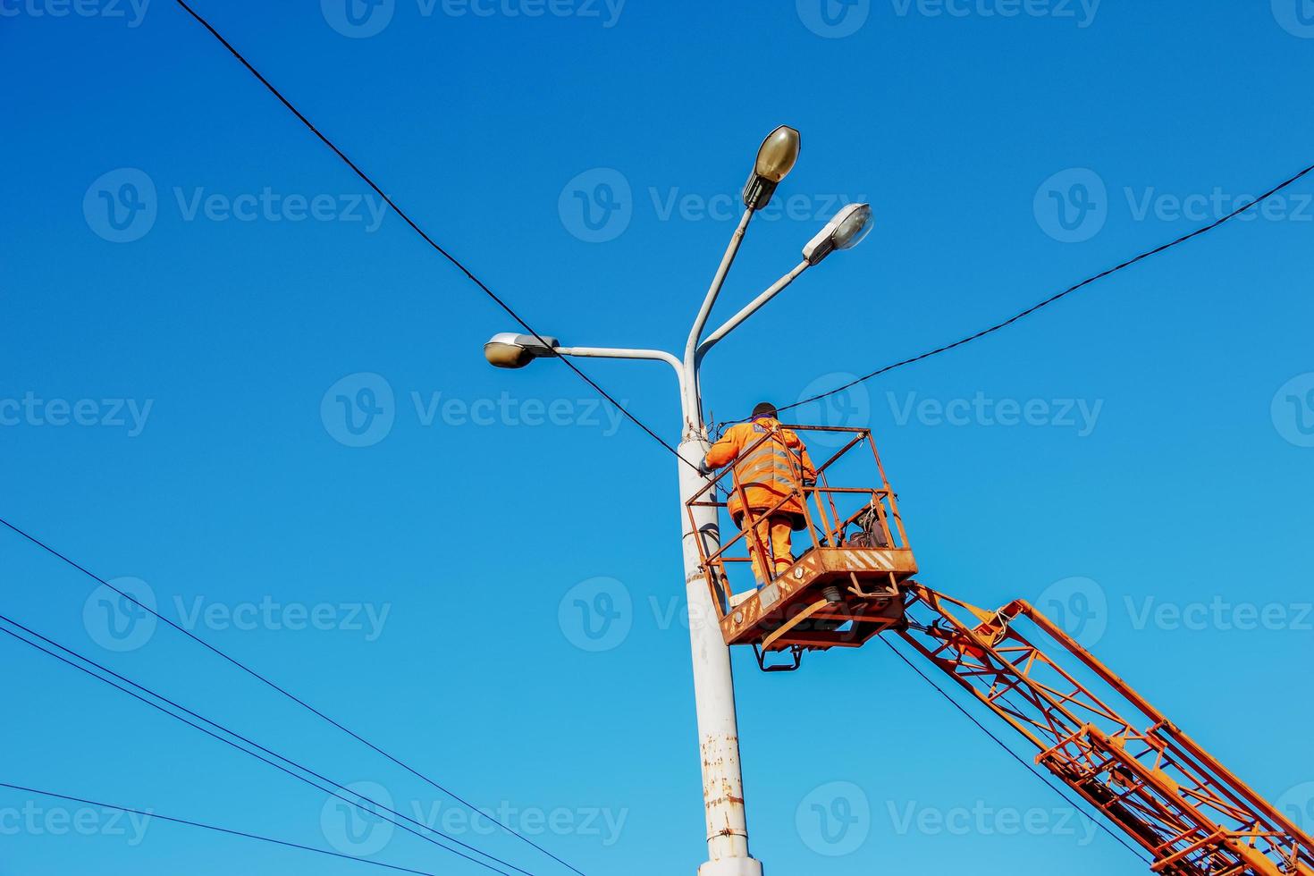 A municipal worker in protective equipment performs hazardous work to eliminate an interruption in the power grid. A worker repairs a street lamp from an aerial platform basket. photo