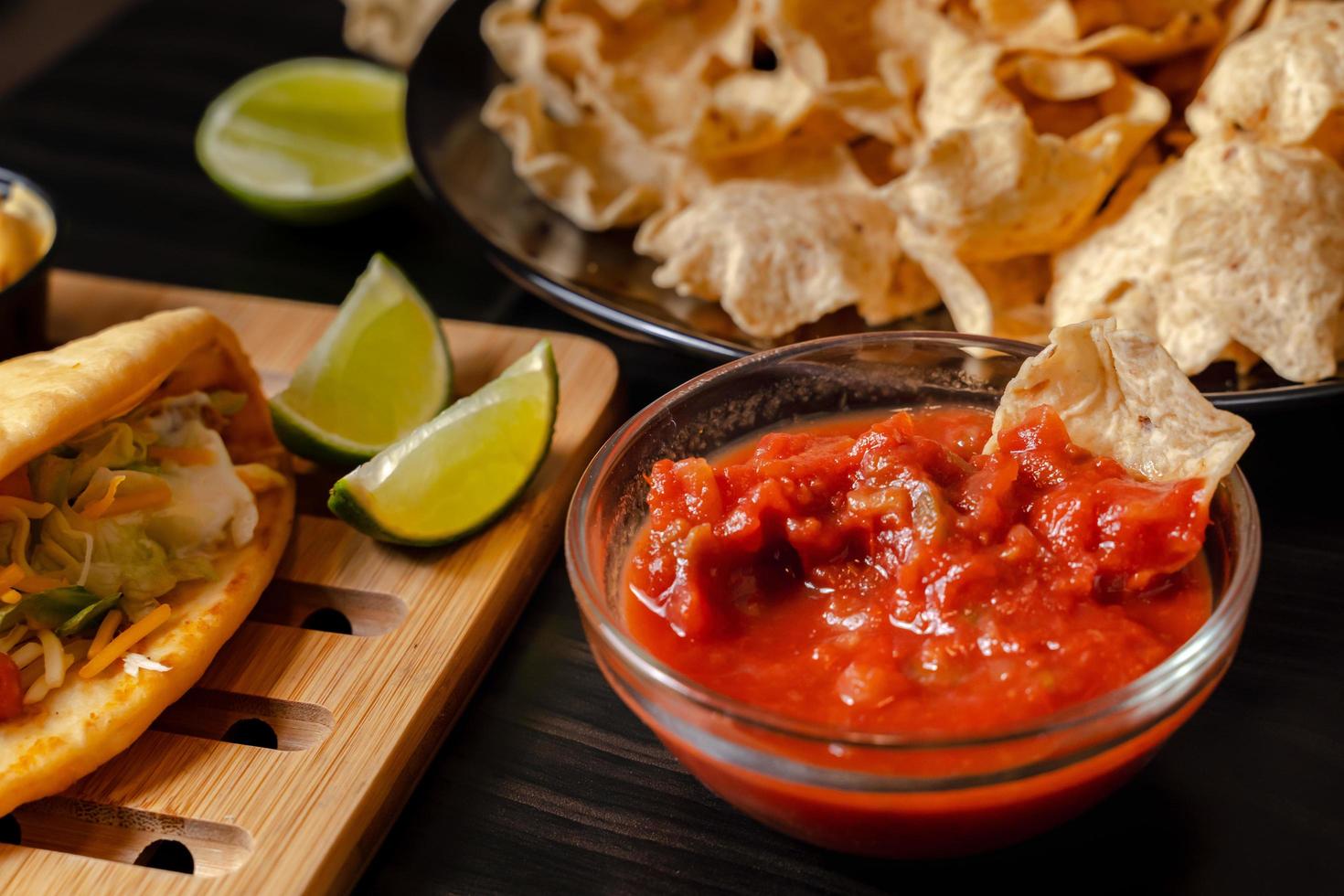 cierre la mano con salsa de tomate con chip dip con plato con taco, chips de nachos y salsa de tomate. nachos picantes sobre fondo de madera rústica foto