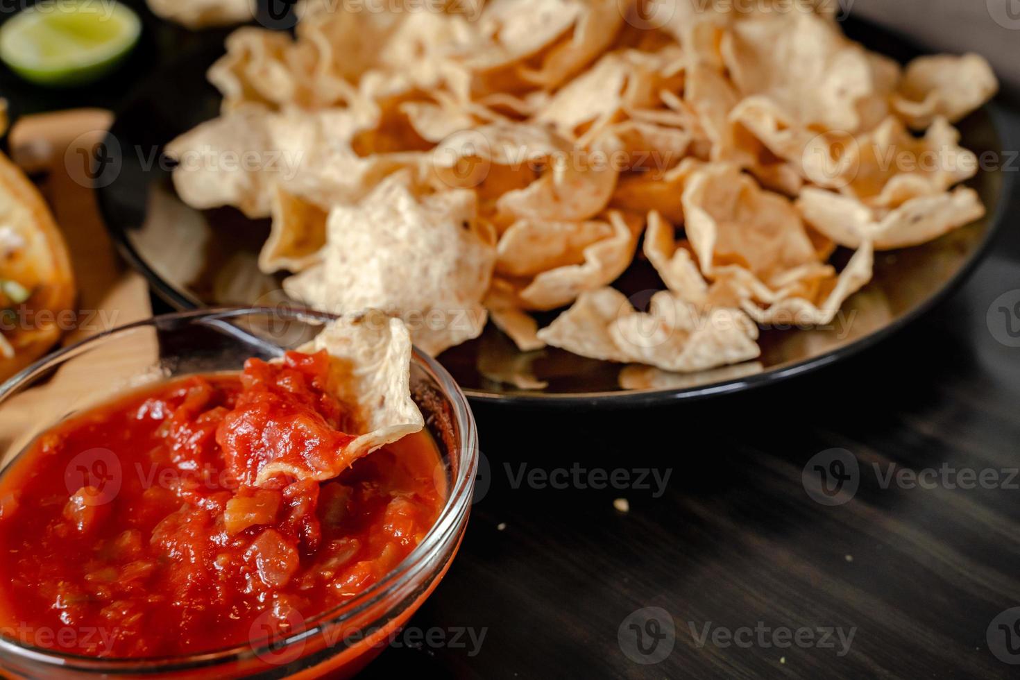 cierre la mano con salsa de tomate con chip dip con plato con taco, chips de nachos y salsa de tomate. nachos picantes sobre fondo de madera rústica foto