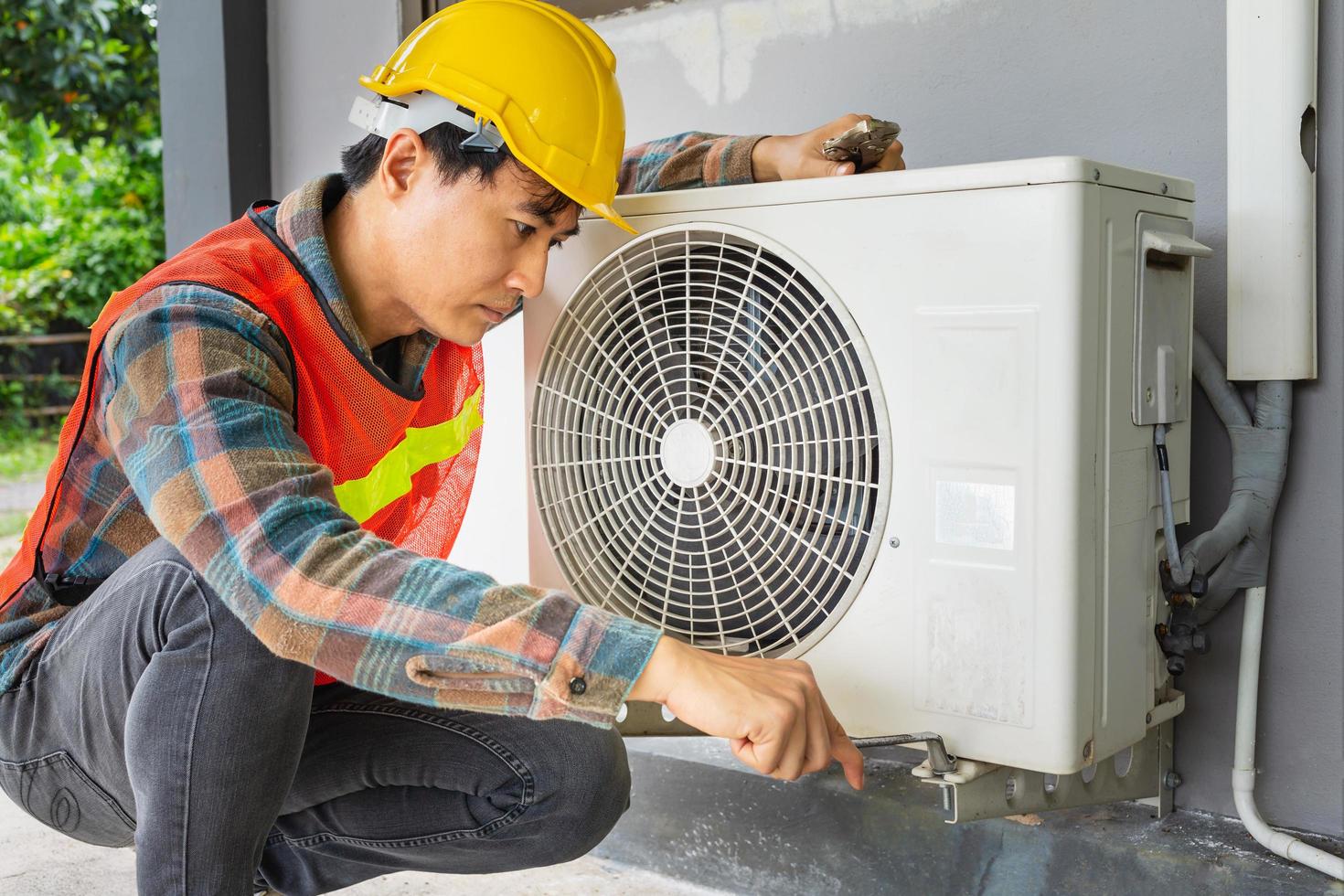 el técnico de aire acondicionado usa una llave para apretar la tuerca del compresor de aire. un joven reparador asiático revisando una unidad de aire acondicionado exterior. foto