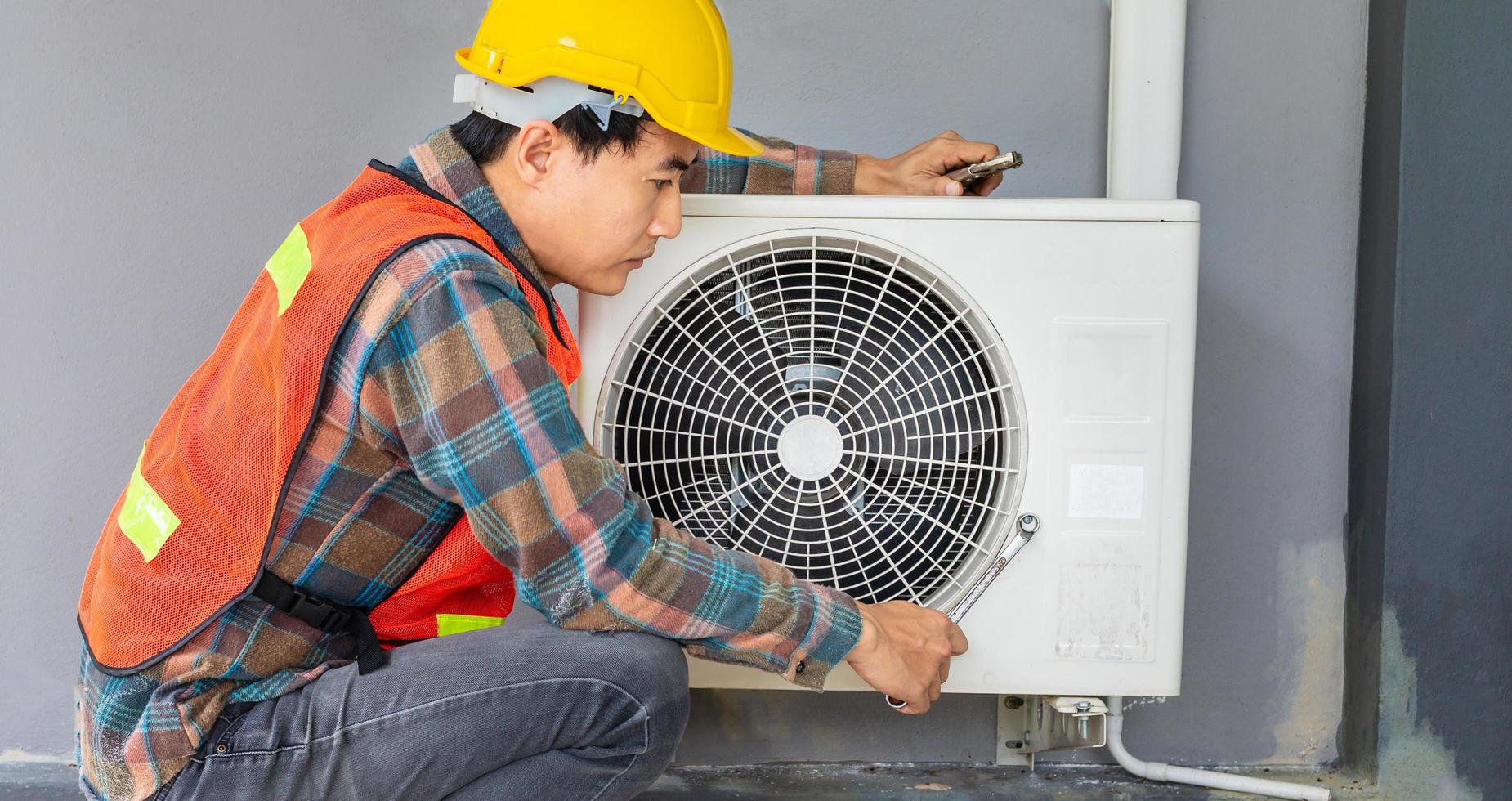 The air conditioner technician Uses a wrench to tighten the nut of the air compressor. Young Asian man repairman checking an outside air conditioner unit. photo