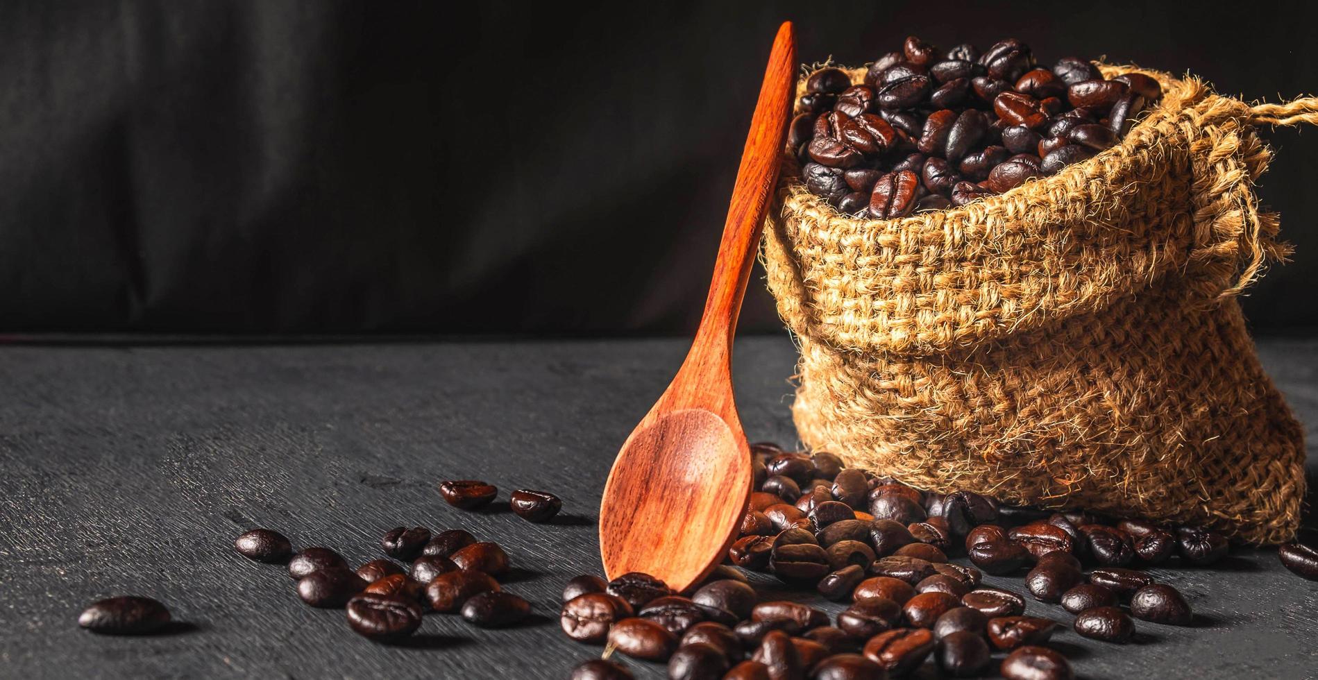 producto granos de café tostados marrones en una bolsa de saco marrón con cuchara de madera sobre fondo de madera negra foto