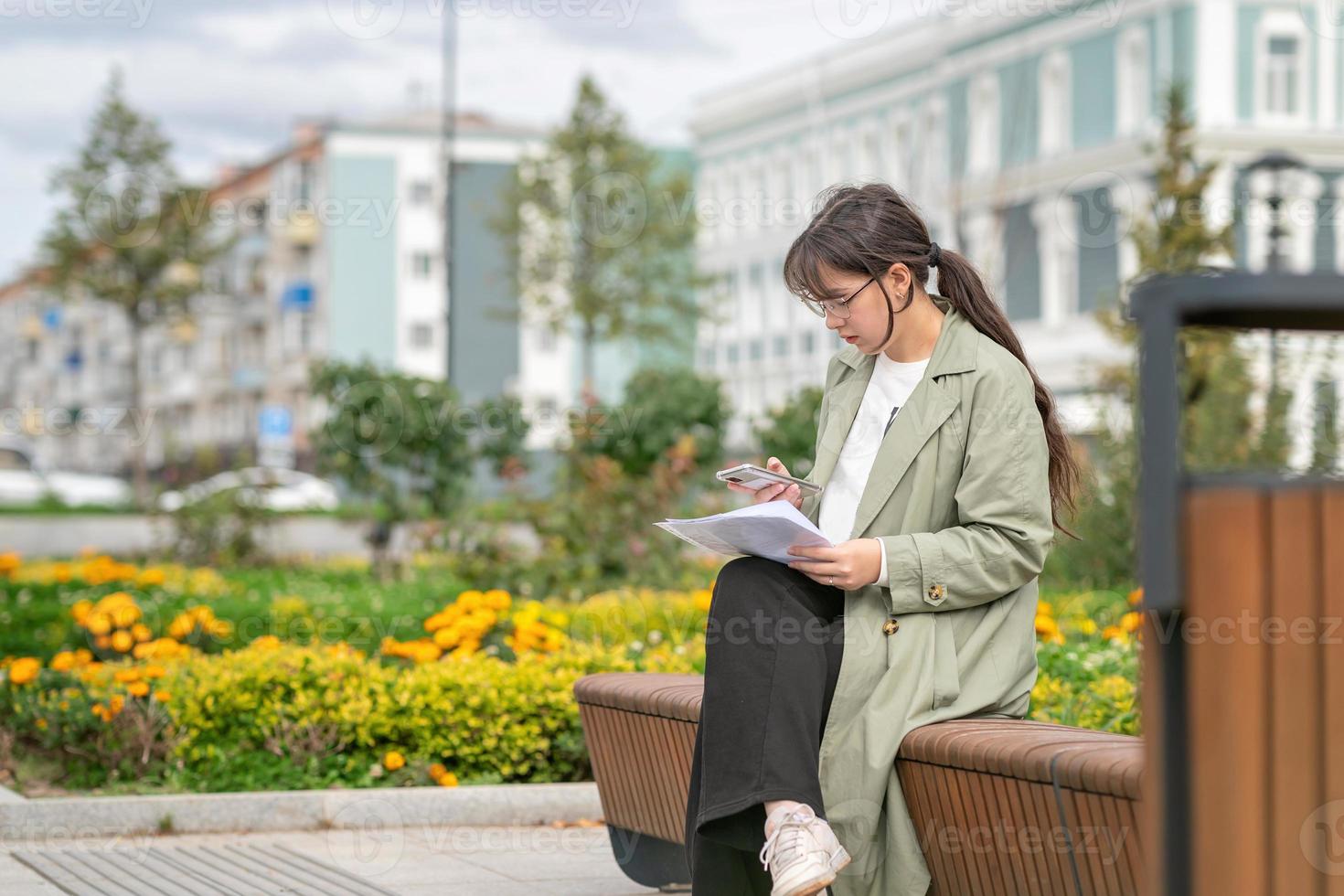 una niña sentada en un banco en la calle con documentos en las manos y usando su teléfono foto