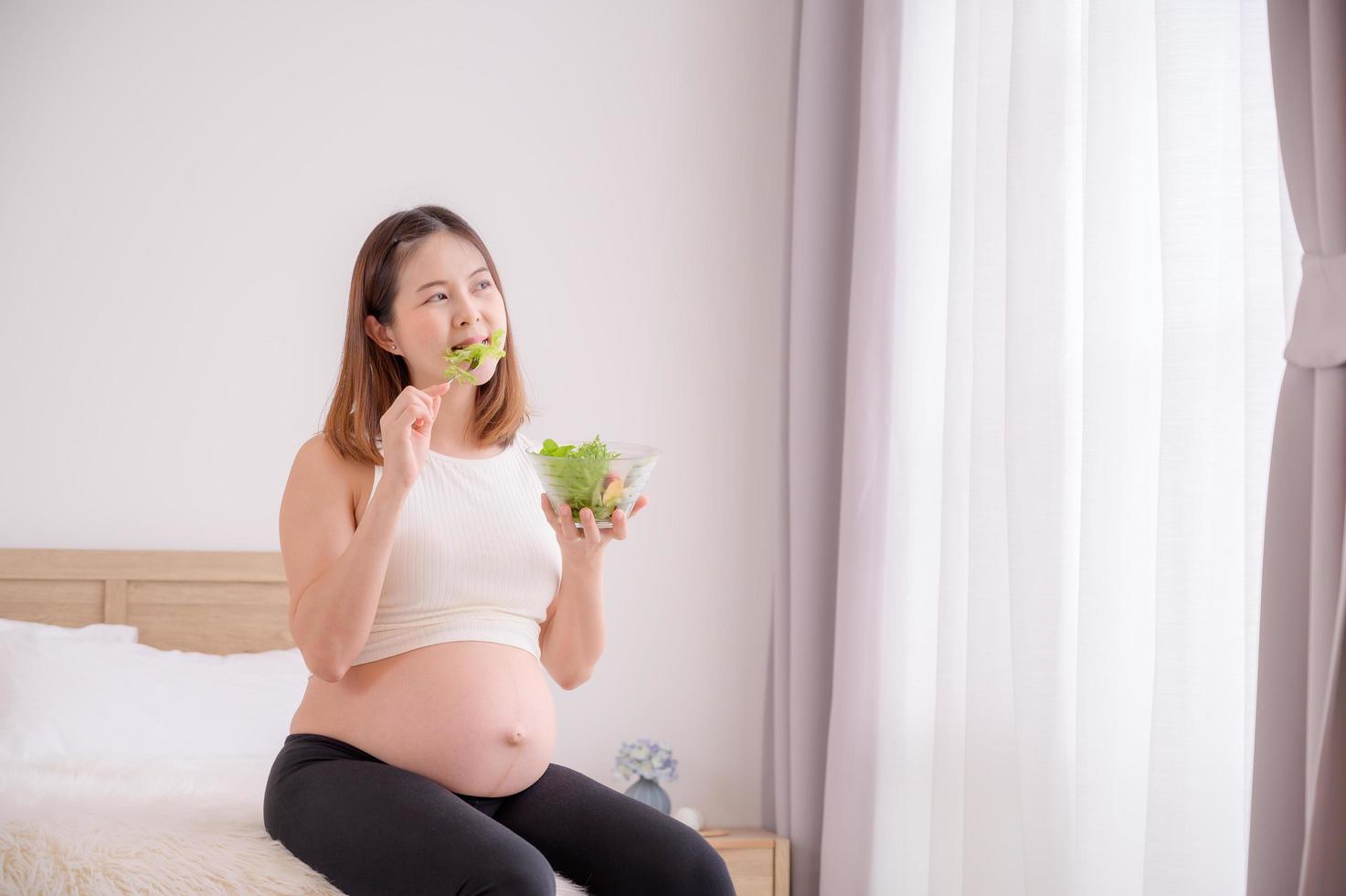una mujer asiática se sienta en su dormitorio comiendo ensaladas orgánicas saludables mientras está embarazada hasta el momento del nacimiento de su bebé foto