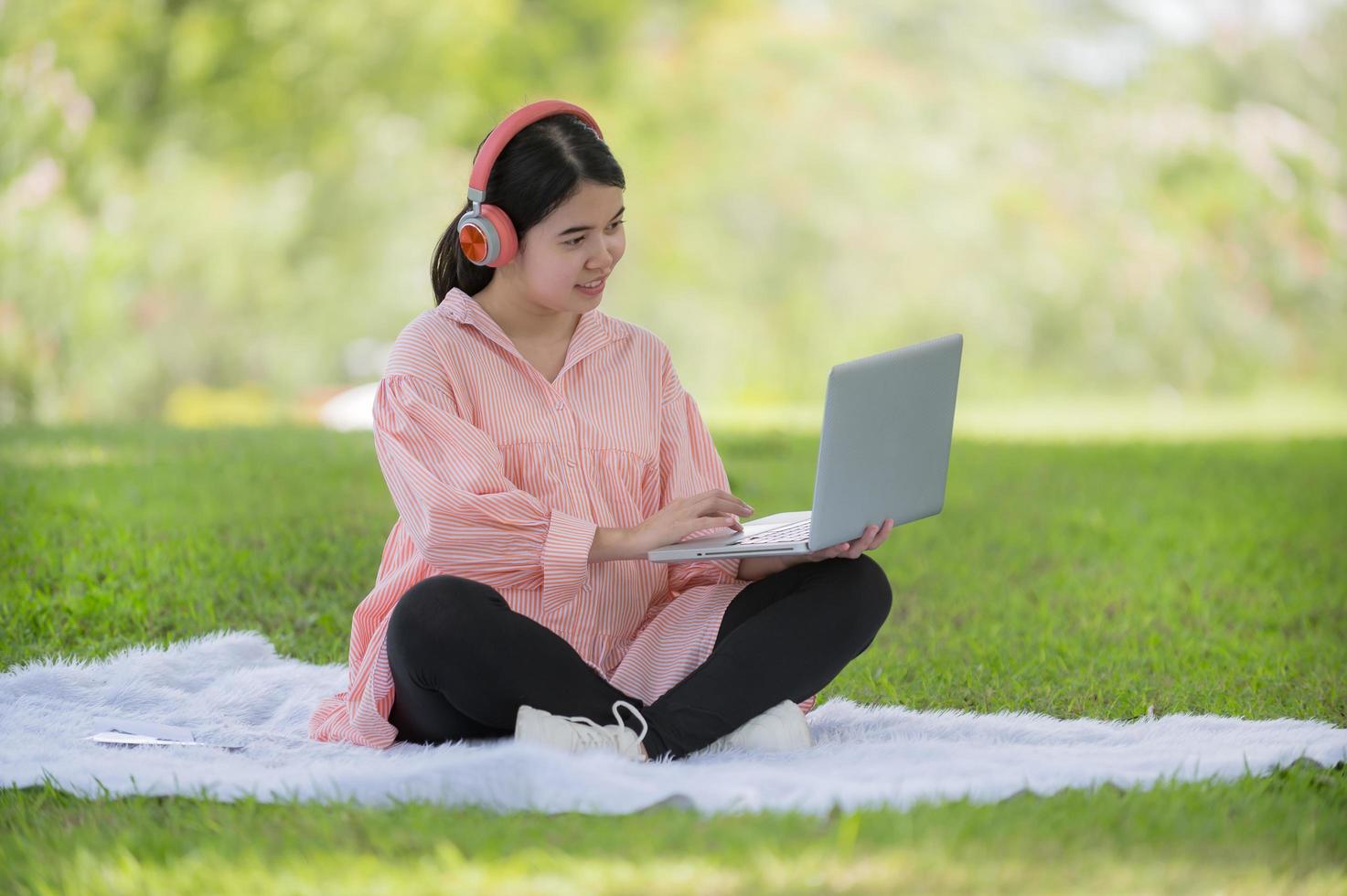 A pregnant Asian woman sits in the garden and puts her headphones on, searching for information about raising an upcoming baby photo