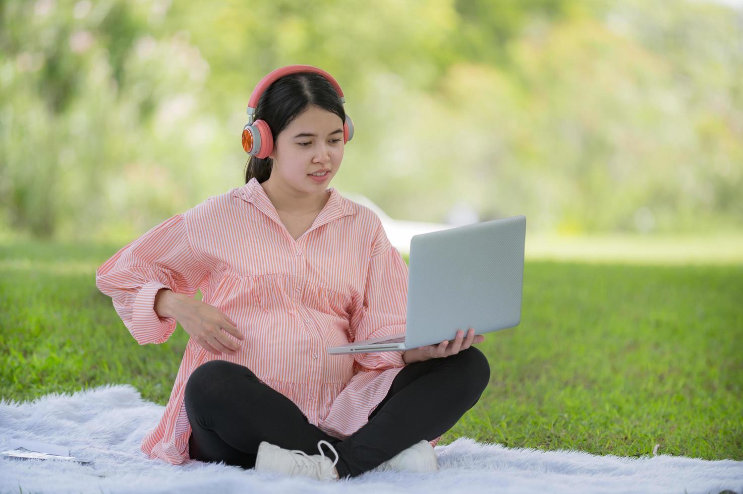 A pregnant Asian woman sits in the garden and puts her headphones on, searching for information about raising an upcoming baby photo