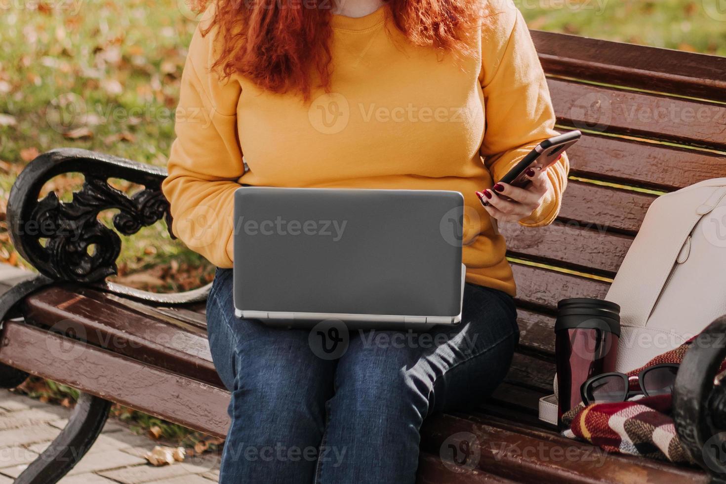 Young red-haired woman freelancer works in park using laptop. Modern businesswoman talking on phone. Online work on Internet. photo